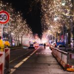 Visitors walk through a German Christmas market in the evening, with festive lights, crowded stalls and visible security presence.