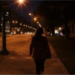 Woman walking alone on a quiet, dimly lit city street at night under streetlights